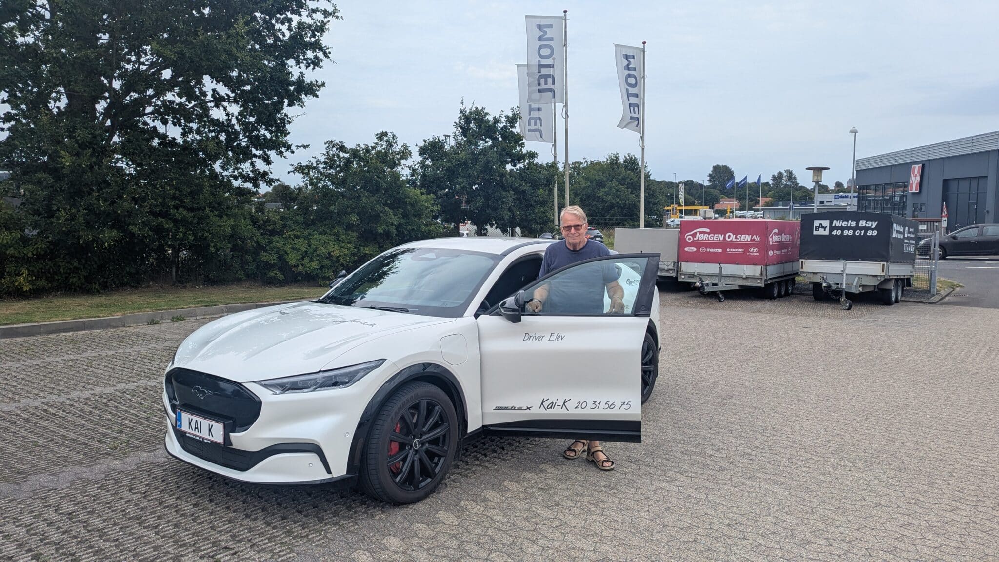 en kørelærer, der står ved en moderne skolebil parkeret i Viborg. Bildøren er åben, og bilen er tydeligt markeret som elevbil. I baggrunden ses trailere, hvilket understøtter undervisning i både almindeligt Kørekort B og udvidelser. Situationen signalerer praktisk køreundervisning, hvor eleven er ved at starte eller afslutte en køretime med fokus på tryghed og rutine i trafikken.